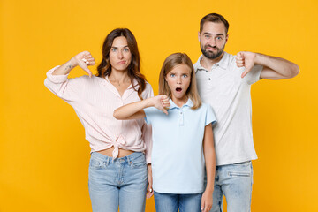Obraz premium Shocked displeased young parents mom dad with child kid daughter teen girl in basic t-shirts showing thumbs down isolated on yellow background studio portrait. Family day parenthood childhood concept.