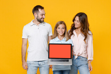 Fototapeta na wymiar Smiling young parents mom dad with child kid daughter teen girl in basic t-shirts hold laptop pc computer with blank empty screen mock up copy space isolated on yellow background. Family day concept.