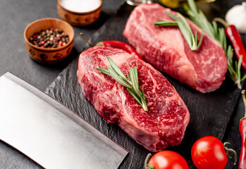 Two raw marbled beef ribeye steaks with spices on a stone background