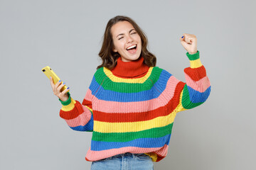 Happy young brunette woman in casual colorful knitted sweater using mobile cell phone typing sms message doing winner gesture keeping eyes closed isolated on grey colour background, studio portrait.