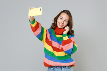 Smiling pretty beautiful attractive young brunette woman 20s wearing casual colorful knitted sweater standing doing selfie shot on mobile phone isolated on grey colour background, studio portrait.
