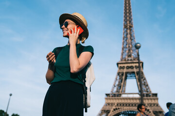 Fototapeta na wymiar Smiling female tourist in trendy apparel enjoying cellphone conversation in roaming for discussing solo vacations in France, happy woman in sunglasses laughing during smartphone calling via app