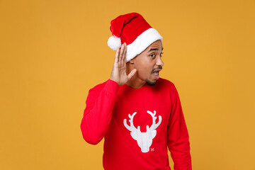 Curious young Santa african american man in red sweater Christmas hat try to hear you overhear listeni intently isolated on yellow background studio. Happy New Year celebration merry holiday concept.