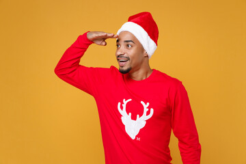 Excited young Santa african american man in Christmas hat holding hand at forehead looking far away distance isolated on yellow background studio portrait. New Year celebration merry holiday concept.