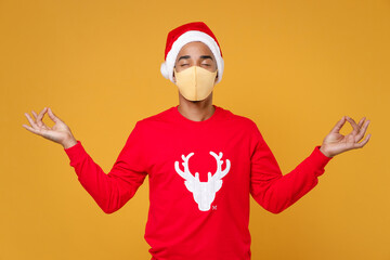Fototapeta na wymiar Santa african american man in Christmas hat mask to safe from coronavirus virus hold hands in yoga gesture relaxing meditating isolated on yellow background Happy New Year celebration holiday concept.