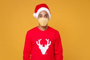 Fototapeta na wymiar Young Santa african american man wearing red sweater Christmas hat face mask safe from coronavirus virus covid-19 isolated on yellow background studio portrait. New Year celebration holiday concept.