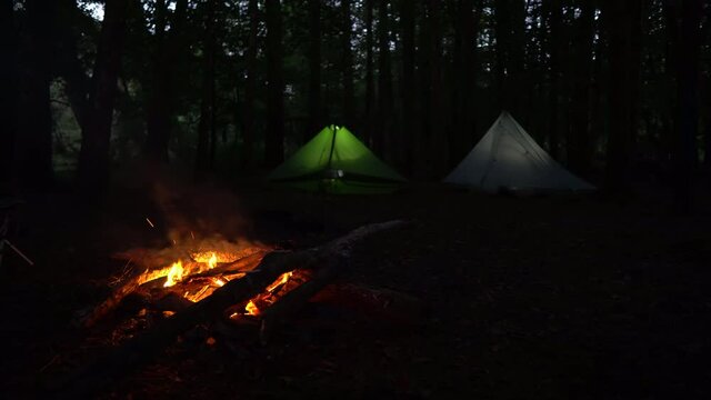 Two glowing tents pitched in forest at night with campfire burning, dolly slide