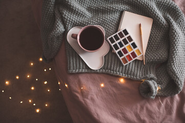 Cup of fresh coffee on plate with palette of paints, sketch book and knitted sweater over glowing...