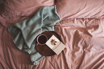 Cup of fresh coffee with open sketch book with fallen autumn leaf on wooden tray and knitted sweater in bed close up. Good morning.