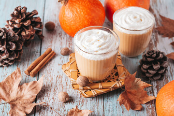 Pumpkin pie smoothie on worn wooden table with cedar cones and dry leaves. Autumn still life with pumpkin smoothie decorated with whipped cream closeup