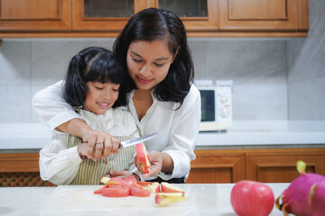 Asian mother is teaching her little daughter cutting apple in the kitchen at home. 