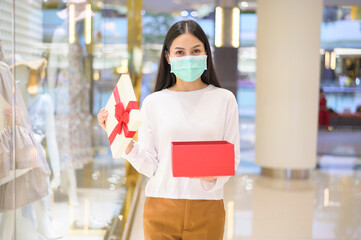 Woman wearing protective mask holding a gift box in shopping mall, shopping under Covid-19 pandemic, thanksgiving and Christmas concept.