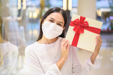 Woman wearing protective mask holding a gift box in shopping mall, shopping under Covid-19 pandemic, thanksgiving and Christmas concept.