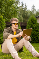 Smart handsome guy in casual clothes and headphones holding tablet in hands watching webinar, listening podcast, siting on grass in park resting working on digital tablet. Online business, e-learning