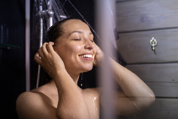 Fototapeta na wymiar Side view of happy beautiful woman washes body and hair under stream of hot water in bathroom. Lifestyle, beauty, personal care, relaxing time, young smiling lady enjoying showering, copy space