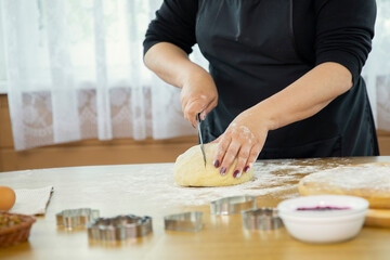 Obraz na płótnie Canvas Caucasian housewife cuts off a piece of raw dough with a knife to make dessert. Cooking festive gingerbread cookies. Wooden kitchen interior.