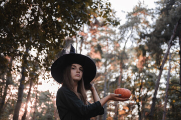 Young cute beautiful woman in dark dress and witch's hat holds an orange pumpkin in her hands....