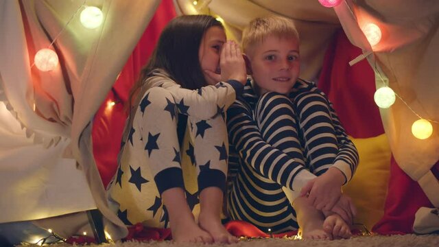 Happy little brother and sister in pajamas telling secrets playing in tent with party lights