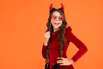 funny child in witch costume hat for Halloween with party glasses and devil horns on orange background, halloween party