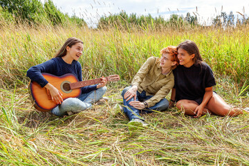 Naklejka na ściany i meble Summer holidays vacation music happy people concept. Group of three friends boy and two girls with guitar singing song having fun together outdoors. Picnic with friends on road trip in nature.