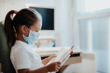 Quarantine training. child little girl studies at home, reading a book by the window, wear protective masks. Distance learning online education.     