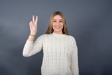 Beautiful young blonde woman wearing white sweater standing against gray background, pointing with fingers number two.