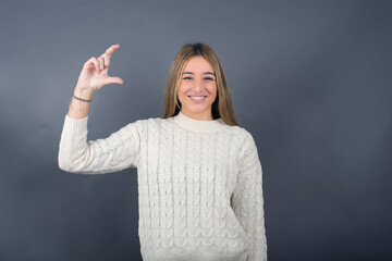 Beautiful young blonde woman wearing white sweater standing against gray background, 