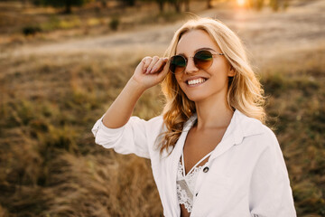 Young happy woman with blond hair posing outdoors, wearing round sunglasses, smiling.