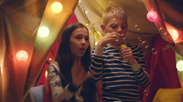 Cute boy and girl having fun in tent with lights blowing soap bubbles