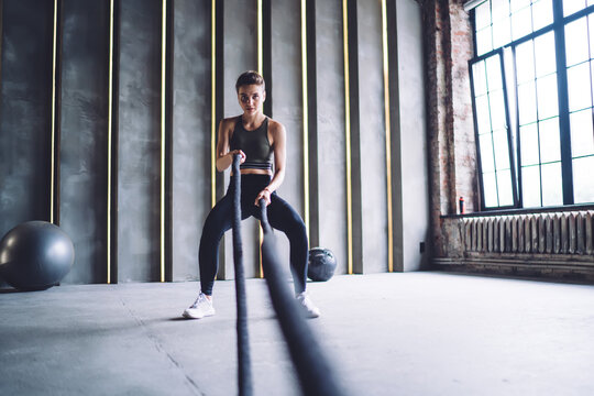 Confident caucasian female in active wear using rope for slimming and working on arms muscles in gym, serious trong professional sports women having hard cardio workout for training strength