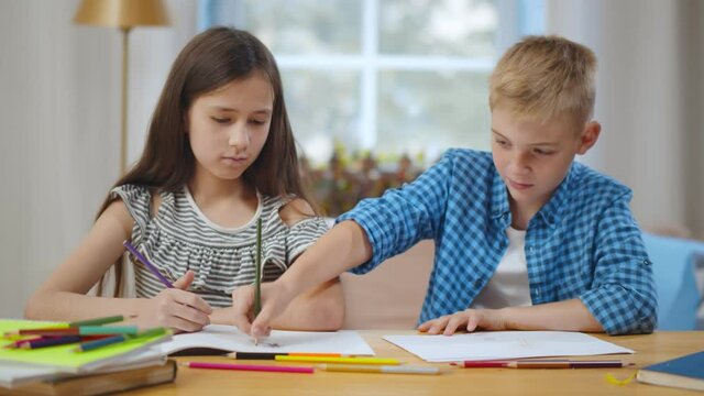 School boy and girl sitting at table and drawing on each other paper with colorful pencils