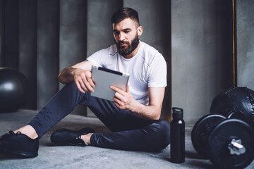 Handsome caucasian bearded male reading information from social networks using modern smartphone, sportsman checking results of workout on application on mobile phone sitting in gym training
