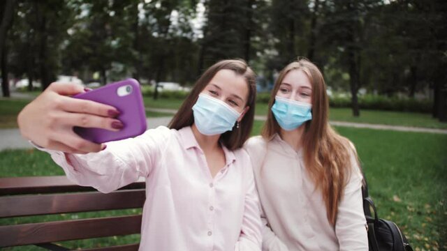 Two young smiling hipster women girls taking a photos selfie on smartphone. Girls students wearing medical face mask making video calling. Female showing positive face emotions.