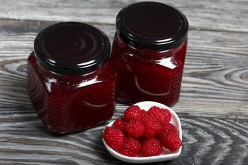Raspberry jam in glass jars. Nearby are raspberries in a saucer. On wooden boards with a beautiful texture.