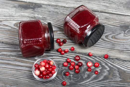 Cranberry jam in glass jars. Nearby berries are in a container and scattered over the surface. On wooden boards with a beautiful texture.