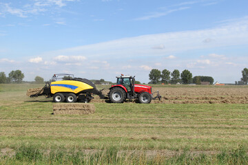 a tractor with a big baler is making straw bales in summer in a typical dutch landscape with a green meadow and a blue sky