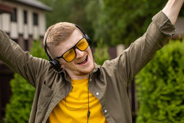 young bearded guy in yellow glasses casually dressed listening to music in headphones and joyfully singing with his hands up on green backdrop.