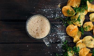Homemade potato chips with parsley and glass with dark beer on dark rustic wooden table. Tasty fast food. Free copy space for text. Background of the Oktoberfest. Top view. Flat lay