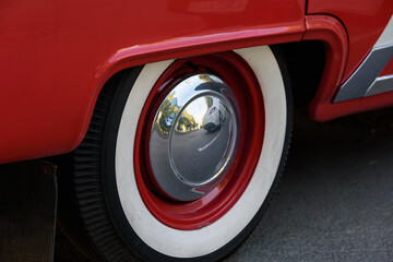 Fototapeta na wymiar Closeup photo of the rear wheel of a magnificent retro car. The wheel has a red stamped disc in the body color, a large chrome hood and white wall.