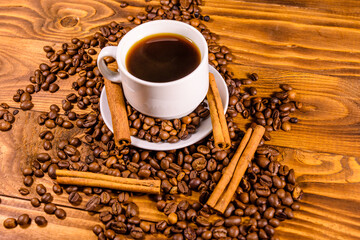 Cup of hot coffee, cinnamon sticks and scattered coffee beans on wooden table