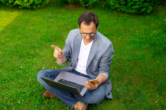 Joyful And Focused Unshaven Mature European 40s Businessman Sits On Grass On Shady Patch With Laptop And Points His Finger At Copy Space. Holiday Work Outdoors