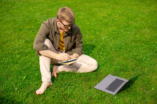 Young millenial guy sitting on grass in park, watching video online distance webinar, training courses, makes notes in notebook. Smart freelancer works, stident learning. Vacation lifestyle outdoors