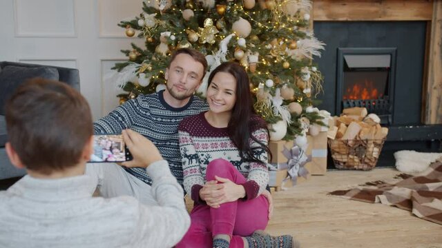 Man and woman are posing near Christmas tree while boy taking pictures with smartphone using modern device at home. People and holidays concept.