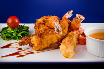 Deep-fried tiger prawns in batter served with sweet and sour sauce, nori seaweed and cherry tomatoes on a rectangular plate.