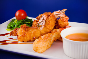 Deep-fried tiger prawns in batter served with sweet and sour sauce, nori seaweed and cherry tomatoes on a rectangular plate.