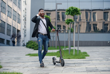 Smiling businessman with electric scooter standing near modern business building talking on phone.