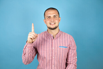 Russian business man wearing a red shirt over blue background showing and pointing up with fingers number one while smiling confident and happy.