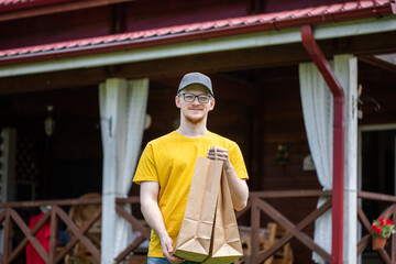 Young handsome delivery man in yellow t-shirt holds biodegradable paper bags in front of country house outdoors, Express delivery service concept, door to door, e-commerce, shipping, online shopping