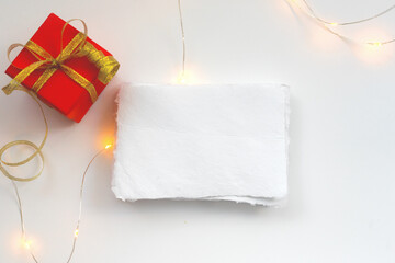 Red gift box with a gold ribbon stands on a white table surface with a sheet of paper with place for text. The concept of greeting cards for the New Year.