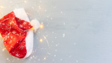 The Santa Claus hat lies on a wooden blue table in the light of a festive garland and snowflakes.